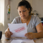 Mulher analisando uma carta de benefício negado em casa, com expressão preocupada, em uma cozinha iluminada por luz natural.